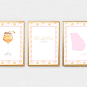 May include: Three framed art prints with gold frames. The prints feature a watercolor cocktail, the text "ATLANTA Georgia", and a pink outline of the state of Georgia. Each print has a white background with a pink and yellow floral border.