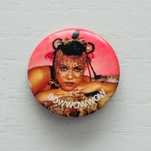 Puede incluir: Botón redondo con un retrato de una persona con pintura facial y el texto "BOW WOW WOW" en una fuente estilizada. El botón tiene un fondo rosa y una superficie blanca.