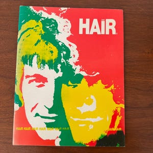 Pode incluir: Uma capa de revista vintage com a palavra "HAIR" em branco sobre um fundo vermelho. A capa apresenta uma ilustração estilizada de dois rostos com cabelo verde, amarelo e branco contra um fundo vermelho. As palavras "HAIR HAIR HAIR HAIR HAIR HAIR HAIR HAIR" são impressas na parte inferior.