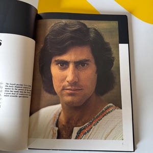 May include: A portrait of a man with dark hair and a white shirt with orange and blue trim. The image is from a book, with text visible on the left page. The man is looking directly at the viewer.