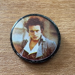 Puede incluir: Un botón redondo negro con una imagen vintage de un hombre con cabello castaño y una camisa a rayas. La imagen tiene un aspecto envejecido, sobre un fondo marrón y beige.