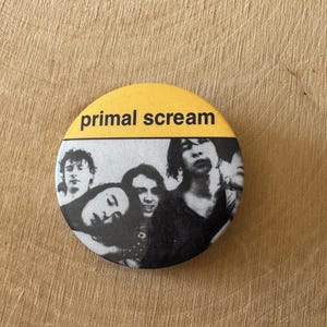Könnte beinhalten: Ein runder Button mit dem Text "primal scream" in Schwarz auf gelbem Hintergrund. Der untere Teil des Buttons zeigt ein Schwarz-Weiß-Bild einer Band. Der Button liegt auf einer hellbraunen Holzoberfläche.