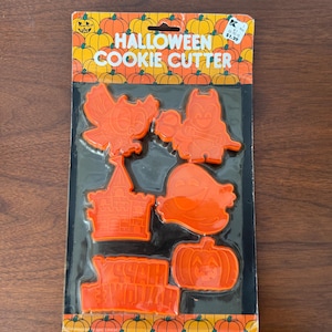 May include: Orange Halloween cookie cutters in a package. The cutters are shaped like a bat, ghost, pumpkin, castle, and a leaf. The package has the words "Halloween Cookie Cutter" and a pumpkin design.