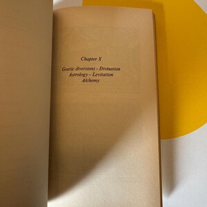 May include: Open book with text on a tan page. The text reads "Chapter X" followed by "Goetic diversions - Divination Astrology - Levitation Alchemy". The book is open on a white and yellow patterned surface.