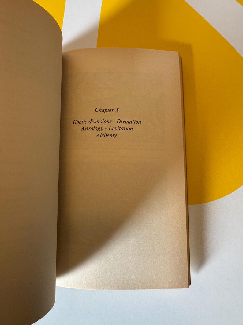 May include: Open book with text on a tan page. The text reads "Chapter X" followed by "Goetic diversions - Divination Astrology - Levitation Alchemy". The book is open on a white and yellow patterned surface.