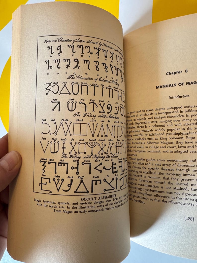 May include: Open book with pages displaying occult alphabets and symbols. The left page features various esoteric characters and writings, while the right page contains text about witchcraft and magic. The book is from the early nineteenth century.