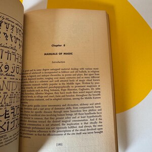 May include: An open book with text on aged paper, featuring the title "MANUALS OF MAGIC" under Chapter 8. The book's pages are cream-colored, with black text. The background includes a yellow and white angled surface.