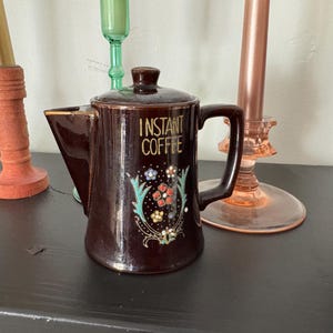 May include: A brown ceramic coffee pot with a gold and green floral design. The pot has a handle and a spout and says "Instant Coffee" on the front.