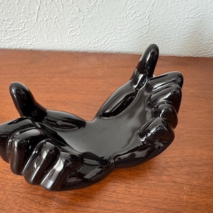 Pode incluir: Uma tigela de cerâmica preta brilhante em forma de mão. A tigela é esculpida para se assemelhar a uma mão aberta, com os dedos e a palma formando uma tigela rasa. A superfície reflete a luz, destacando as curvas e contornos do design. Esta peça decorativa é adequada para guardar pequenos objetos.