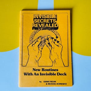 Op de afbeelding: Een geel boek met de titel "INVISIBLE SECRETS REVEALED" met een illustratie van handen die speelkaarten vasthouden. Het boek luidt ook "New Routines With An Invisible Deck" door Eddie Fields & Michael Schwartz.