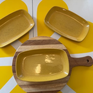 May include: Three rectangular mustard yellow ceramic dishes with a subtle speckled finish. Two dishes are positioned above a round wooden serving board with a handle, while the third dish rests directly on the center of the wooden board.