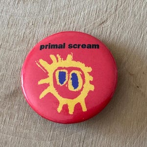 Könnte beinhalten: Ein roter runder Button mit dem schwarzen Schriftzug "primal scream". Darunter befindet sich ein gelbes sonnenähnliches Design mit blauen Augen. Der Button ist rund und scheint aus Metall zu sein.