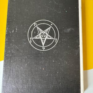 May include: A black book with a white pentagram symbol on the cover. The pentagram features a star within a circle, with a goat-like face in the center. The book has a textured surface and a white spine.