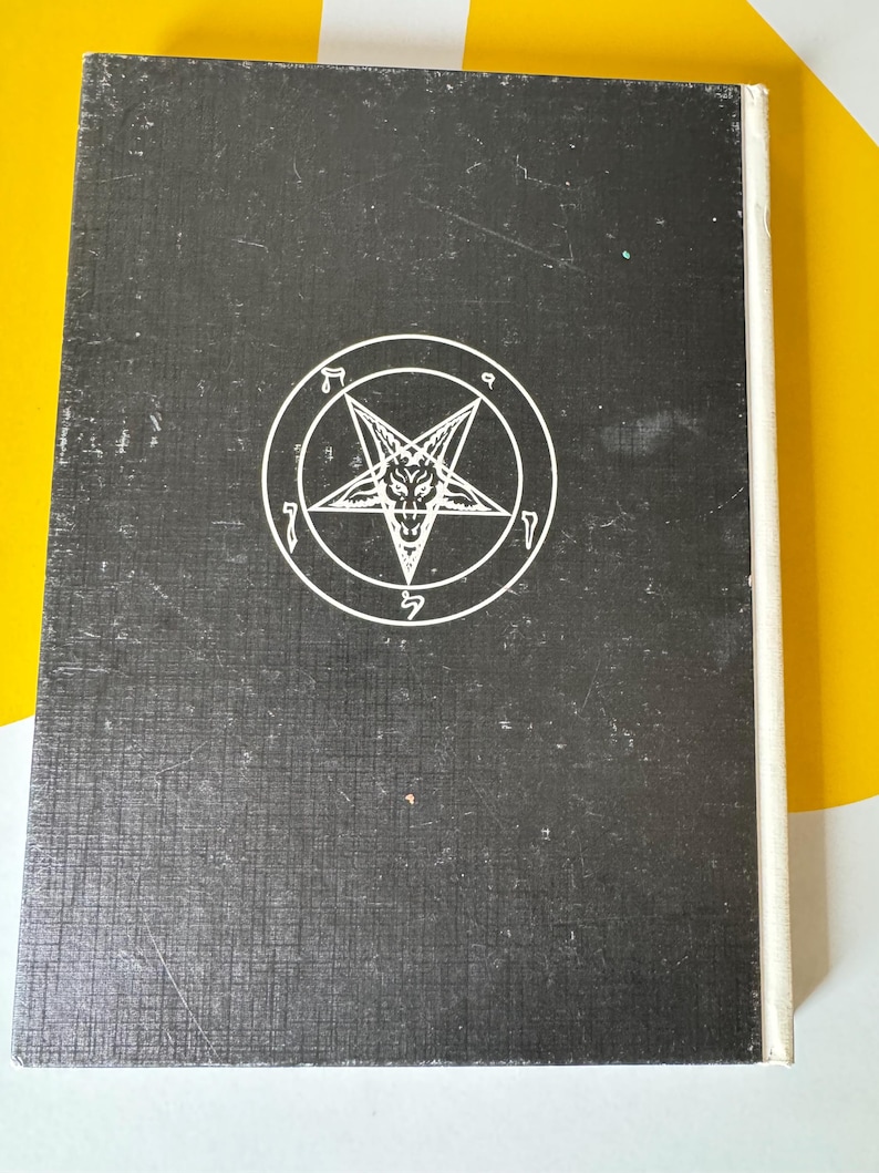 May include: A black book with a white pentagram symbol on the cover. The pentagram features a star within a circle, with a goat-like face in the center. The book has a textured surface and a white spine.