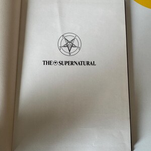 May include: Open book with the title "THE SUPERNATURAL" printed in black. Above the title is a circular emblem with a five-pointed star inside a circle. The book's pages are off-white.