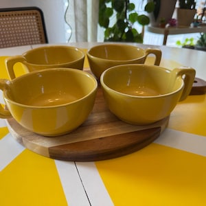 Peut inclure: Quatre bols à soupe en céramique jaune moutarde avec des anses larges et plates, disposés sur un plateau de service rond en bois. Chaque bol présente une finition lisse et brillante ainsi qu'un bassin profond et arrondi.