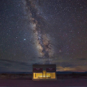 May include: A two-story building with the word "PRADA" on the front, set against a night sky with a bright Milky Way galaxy.