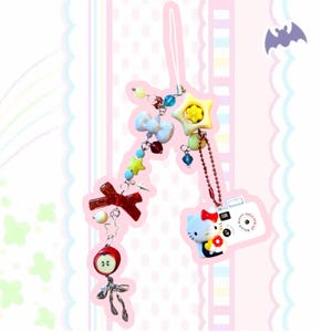 May include: A colorful charm featuring a Hello Kitty camera, bows, stars, and an apple. The charm includes a white string, red chain, and various beads in blue, yellow, and red. The camera has the text "SMILE FOR ME".