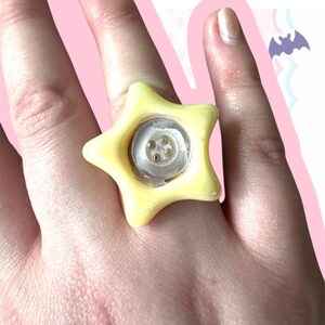 May include: A yellow star-shaped ring with a clear center, featuring a small, round, white button with four holes. The ring is worn on a finger. The ring is a unique piece of jewelry.