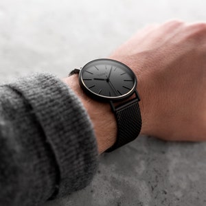 May include: A black wristwatch with a mesh band is displayed. The watch face is black with gold-colored accents and the brand name "Venio" is visible. The watch is on a wrist, with a gray sweater sleeve in the background.
