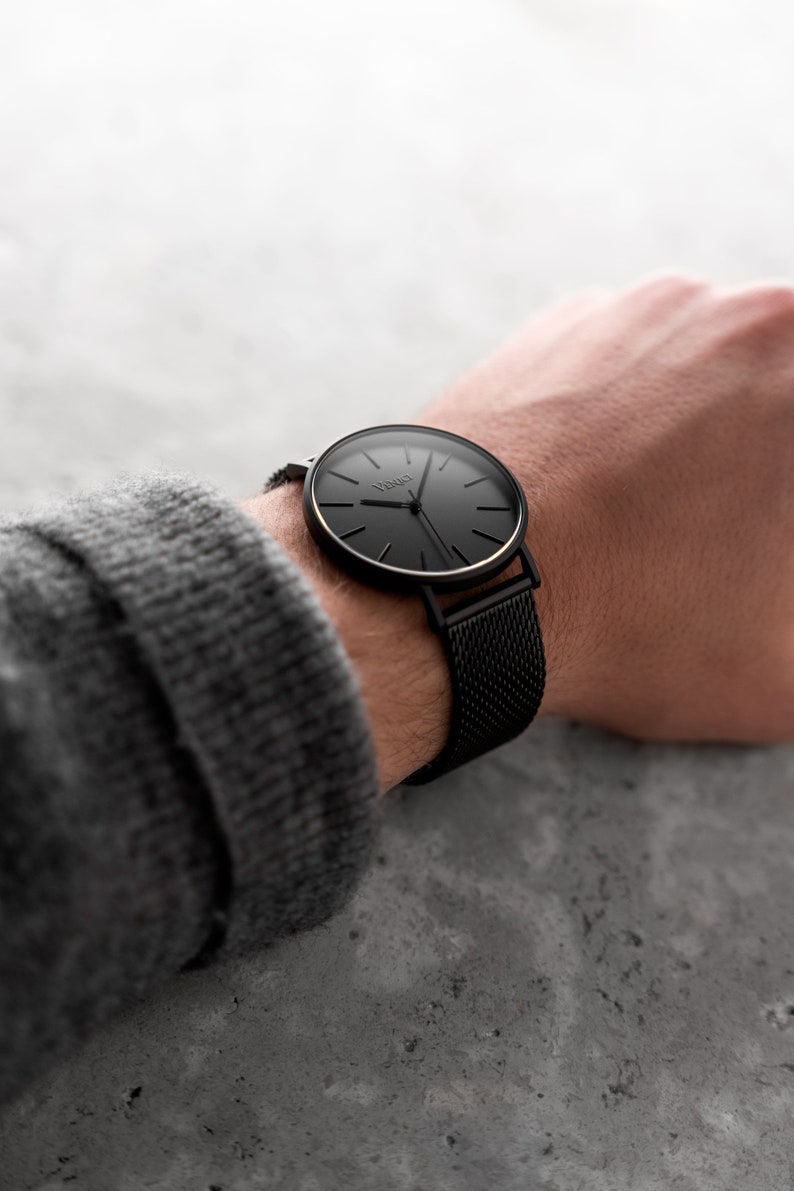 May include: A black wristwatch with a mesh band is displayed. The watch face is black with gold-colored accents and the brand name "Venio" is visible. The watch is on a wrist, with a gray sweater sleeve in the background.