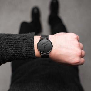 May include: A black wristwatch with a mesh band and a round face. The watch has a minimalist design with black hands and markers. The brand name "Vejar" is printed on the watch face. The watch is worn on a wrist.