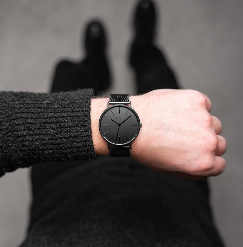 May include: A black wristwatch with a mesh band and a round face. The watch has a minimalist design with black hands and markers. The brand name "Vejar" is printed on the watch face. The watch is worn on a wrist.