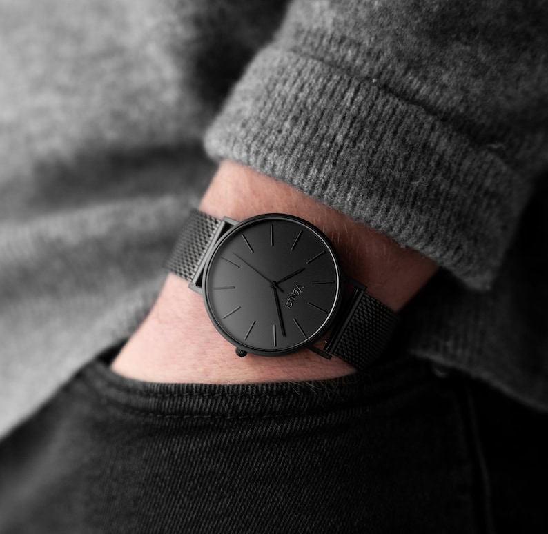 May include: A black wristwatch with a mesh band is displayed. The watch has a minimalist design with a black face and hands. The watch is worn on a wrist, with a gray sweater and black jeans in the background.