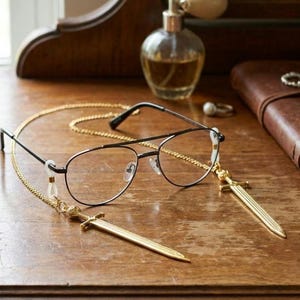 May include: Black eyeglasses with a gold chain and sword-shaped charms rest on a wooden surface. The glasses have a thin black frame and clear lenses. The gold chain is attached to the glasses with small white loops. A gold sword charm hangs from each side.