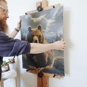 May include: An oil painting of a bear on an easel. The artwork features a detailed depiction of a brown bear against a backdrop of a cloudy sky. The painting is held by a person, showcasing the artwork's size and presentation.