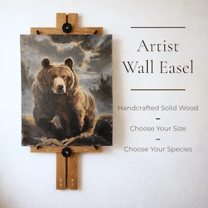 May include: A handcrafted solid wood artist wall easel holding a painting of a bear. The easel is made of light-colored wood and features black hardware. Text on the wall reads "Artist Wall Easel - Handcrafted Solid Wood - Choose Your Size - Choose Your Species."