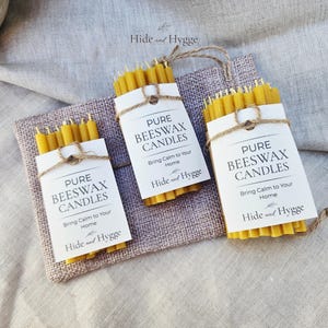 K&ouml;nnte beinhalten: Drei B&uuml;ndel gelber Bienenwachskerzen werden auf einem quadratischen Jutestoff pr&auml;sentiert. Jedes ist mit einem wei&szlig;en Etikett und Bindfaden umwickelt. Die Etiketten lauten "PURE BEESWAX CANDLES" und "Bring Calm to Your Home".
