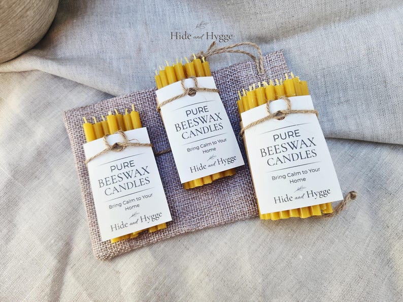 K&ouml;nnte beinhalten: Drei B&uuml;ndel gelber Bienenwachskerzen werden auf einem quadratischen Jutestoff pr&auml;sentiert. Jedes ist mit einem wei&szlig;en Etikett und Bindfaden umwickelt. Die Etiketten lauten "PURE BEESWAX CANDLES" und "Bring Calm to Your Home".