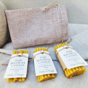 K&ouml;nnte beinhalten: Drei B&uuml;ndel gelber Bienenwachskerzen, jeweils mit Bindfaden und einem wei&szlig;en Etikett mit der Aufschrift "Pure Beeswax Candles" umwickelt. Hinter den Kerzen befindet sich eine Jutetasche mit einem Kordelgriff. Der Text "Bring Calm to Your Home" steht auf den Etiketten.