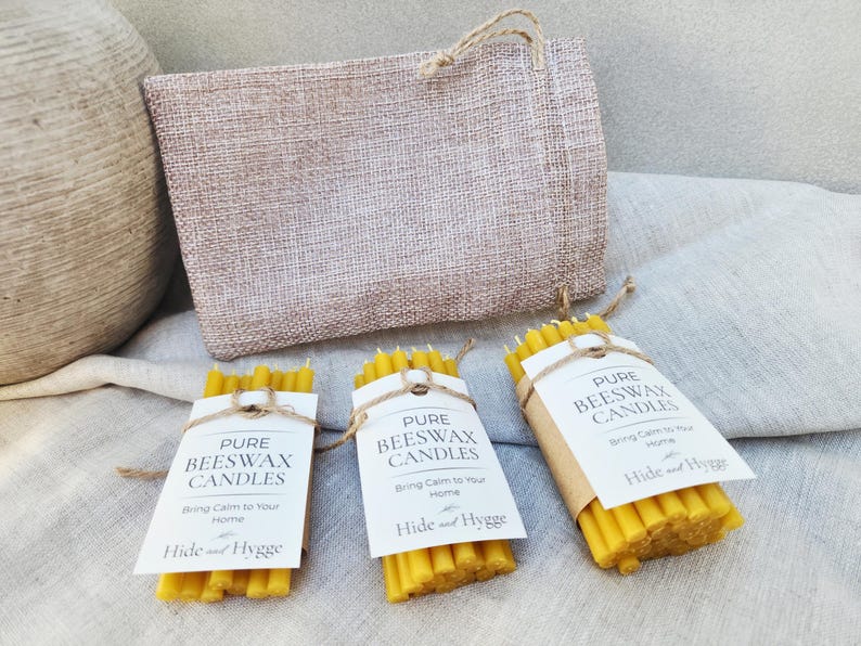 K&ouml;nnte beinhalten: Drei B&uuml;ndel gelber Bienenwachskerzen, jeweils mit Bindfaden und einem wei&szlig;en Etikett mit der Aufschrift "Pure Beeswax Candles" umwickelt. Hinter den Kerzen befindet sich eine Jutetasche mit einem Kordelgriff. Der Text "Bring Calm to Your Home" steht auf den Etiketten.