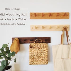 May include: Three solid wood peg rails in oak, maple, and walnut, mounted on a white wall. The top rail is oak, the middle is maple, and the bottom is walnut. A woven basket and a tote bag hang below.