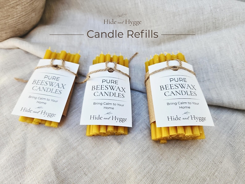 K&ouml;nnte beinhalten: Drei B&uuml;ndel aus reinen Bienenwachs-Kerzen-Nachf&uuml;llungen. Jedes B&uuml;ndel ist mit einer Papiermarke und Bindfaden gebunden. Auf den Etiketten steht "PURE BEESWAX CANDLES" und "Bring Calm to Your Home". Der Markenname "Hide and Hygge" ist ebenfalls sichtbar.