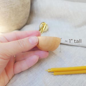 May include: A small, light-colored wooden bowl, approximately 1 inch tall, is held in a person's hand. Yellow candles are in the background. The bowl is empty and appears to be newly made.