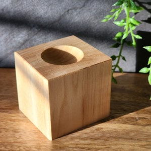 Pode incluir: Um pequeno vaso quadrado de madeira com um centro redondo oco. O vaso é feito de madeira clara e tem um acabamento liso.