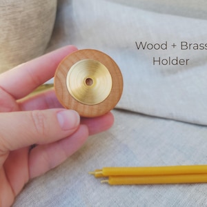 May include: A wooden candle holder with a brass center is held in a hand. The text "Wood + Brass Holder" is visible. Three yellow candles are in the foreground. The holder is approximately 2 inches in diameter.