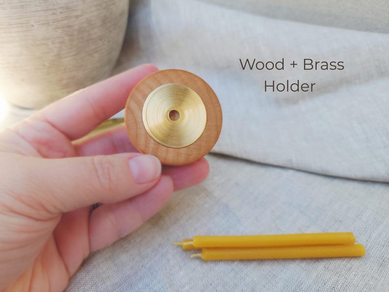 May include: A wooden candle holder with a brass center is held in a hand. The text "Wood + Brass Holder" is visible. Three yellow candles are in the foreground. The holder is approximately 2 inches in diameter.