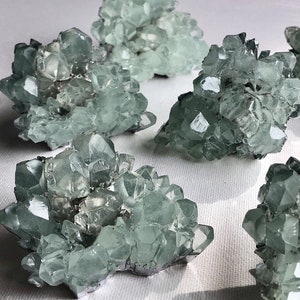 Może przedstawiać: Grupa ośmiu zielonych skupisk kryształów celestynowych. Kryształy są przezroczyste i mają jasnozielony kolor. Skupiska są ułożone w losowym wzorze na białym tle.