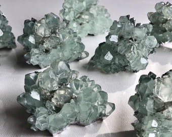 Puede incluir: Un grupo de ocho racimos de cristales de celestina verdes. Los cristales son translúcidos y tienen un color verde claro. Los racimos están dispuestos en un patrón aleatorio sobre una superficie blanca.