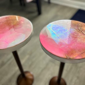 Puede incluir: Dos tapas de mesa redondas de resina rosa y dorada con acentos iridiscentes sobre soportes de madera.