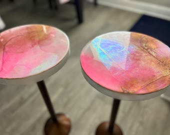 May include: Two round pink and gold resin tabletops with iridescent accents on wooden stands.