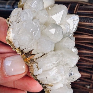 Może przedstawiać: Gromada białych kryształów kwarcu z pozłacanym obrzeżem wokół podstawy. Kryształy są ułożone w skupisku, niektóre z nich skierowane są do góry, a inne do dołu.