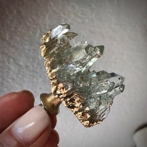 Może przedstawiać: Gromada przezroczystych kryształów kwarcu z złotymi akcentami, zamontowana na mosiężnej podstawie. Kryształ jest naturalną formacją o unikalnym kształcie i fakturze.