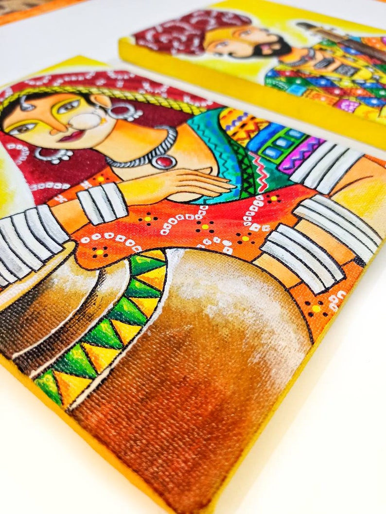 May include: A pair of colorful paintings on canvas depicting a woman and a man in traditional Indian clothing. The woman is wearing a red and yellow sari with a white head covering and a large nose ring. The man is wearing a blue and yellow turban with a red and yellow shirt. Both paintings have a yellow background.