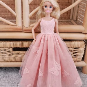 May include: A doll with blonde hair wearing a pink dress with butterfly accents. The dress has a fitted bodice and a full skirt made of tulle. The doll is standing in front of a wicker chair.