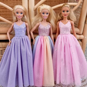 May include: Three fashion dolls with blonde hair and blue eyes, each wearing a different pastel-colored gown. The gowns are lavender, rainbow, and pink, with sequin bodices and full skirts. The dolls are posed in front of a woven background.